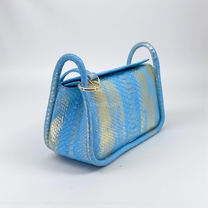 Op de afbeelding: Een lichtblauwe handtas met een slangenpatroon in goud en blauw. De tas heeft twee gevlochten handvatten en een goudkleurige sluiting. De tas is rechthoekig met afgeronde randen.