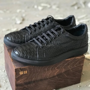Puede incluir: Un par de zapatillas bajas negras con una parte superior texturizada de piel de serpiente, cordones negros y suela de goma negra. El texto AkramReptile es visible en el lateral del calzado, colocado sobre una caja de madera oscura.