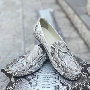 May include: A pair of white, grey, and black snakeskin loafers. The shoes feature a snakeskin pattern and are placed on a snakeskin surface. The loafers have a classic slip-on design.