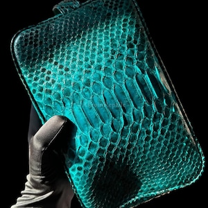 May include: Teal-colored rectangular clutch with a snakeskin pattern. The clutch has a small loop on one end and a textured surface. The color is a vibrant teal, with darker areas creating depth. The overall design is sleek and modern.