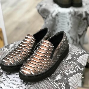 May include: Slip-on shoes with a snakeskin design in brown, bronze, and grey tones. The shoes have a black sole and a rounded toe. The shoes are displayed on a surface with a similar snakeskin pattern.