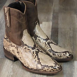 Könnte beinhalten: Ein Paar braune und beige Cowboy-Stiefel mit Schlangenmuster auf der Vorder- und Seite. Die Stiefel haben einen kurzen Schaft, einen Reißverschluss an der Seite und ein Obermaterial aus Leder. Die Stiefel werden auf einer Holzoberfläche präsentiert.