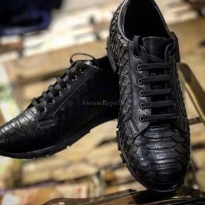 Puede incluir: Un par de zapatillas negras con un patrón texturizado de escamas en el exterior. Los zapatos tienen cordones negros, punta redondeada y el texto AkromReptile es visible en el lateral del zapato situado en el fondo.