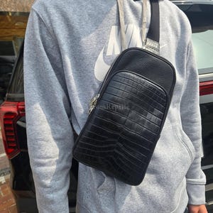 Op de afbeelding: Zwarte crossbodytas met een krokodillenleertextuur. De tas heeft een verticaal ontwerp met een ritssluiting en een zwarte band. De tas wordt over de schouder gedragen.