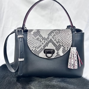 May include: Black handbag with a snakeskin flap and a top handle. The bag features a detachable shoulder strap, a metal clasp, and a decorative tassel. The handle is a dark purple color.