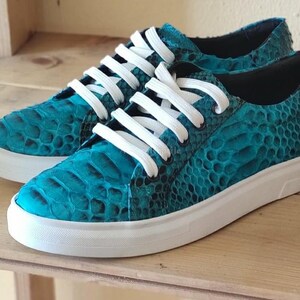 Op de afbeelding: Een paar lage sneakers met een gestructureerd slangenleerpatroon in turkoois en zwart. De schoenen zijn voorzien van witte veters en een effen witte rubberen zool, wat een scherp contrast vormt met het gedetailleerde, schubachtige bovenmateriaal.