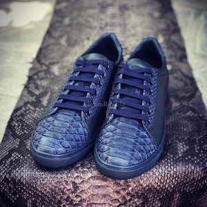 Könnte beinhalten: Ein Paar Sneaker mit niedrigem Schaft, gefertigt aus tiefblauem Leder mit Schlangenledermuster, dunkelblauen Schnürsenkeln und passenden blauen Gummisohlen. Das Design umfasst glatte Lederseitenteile und eine Schuppenstruktur an der Schuhspitze.