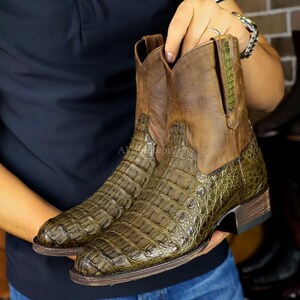 May include: A pair of olive green and brown leather cowboy boots with a crocodile skin texture. The boots have a square toe, a stacked heel, and a pull-on design. The upper part of the boot is brown, while the lower part is olive green.