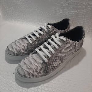 May include: A pair of low-top sneakers featuring a grey and white snakeskin-patterned upper with a textured scale design. The shoes include white laces, a white rubber sole, and a black interior lining, presenting a sleek and detailed aesthetic.