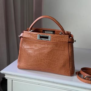 May include: A brown leather handbag with a crocodile skin texture. The bag has a top handle, a silver-coloured rectangular clasp, and a detachable shoulder strap. The brand name "Akron Reptile" is visible on the bag.