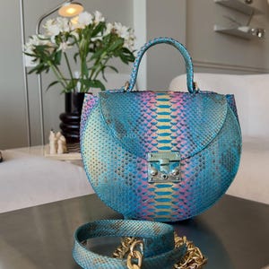 Puede incluir: Un bolso redondo de piel de serpiente azul y rosa, con un asa superior y un cierre plateado. Una correa a juego de piel de serpiente con una cadena dorada está al lado del bolso. El bolso está sobre una superficie plateada.