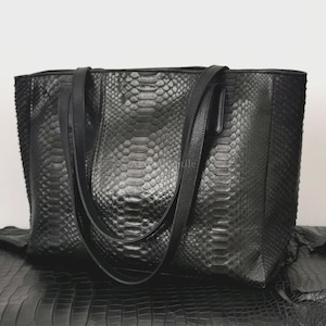 May include: A black tote bag with a snakeskin pattern. The bag has two shoulder straps and a rectangular shape. The bag is made of a textured material and appears to be leather. The bag is sitting on a black surface.