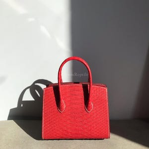 May include: A vibrant red handbag with a textured, reptile-like pattern. The bag features a structured design with a top handle and white stitching details. The bag is a square shape and appears to be made of leather.