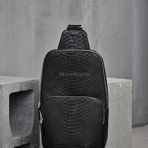 Pode incluir: Bolsa transversal de couro preto texturizado com um bolso frontal e fecho de zíper. A bolsa tem uma alça de ombro e o texto "AkromReptile" é visível na frente.