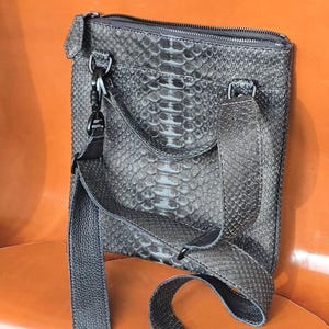 May include: A gray crossbody bag with a snake-skin pattern. The bag has a zipper closure and a long, adjustable strap. The hardware is black. The bag is made of a textured material.