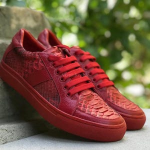 May include: A pair of vibrant red sneakers featuring a textured snakeskin pattern on the toe box and side panels, complemented by smooth leather overlays, matching red laces, and a solid red rubber sole with the text AkromReptile visible.
