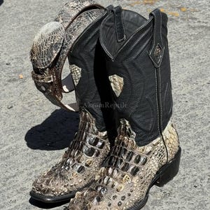 Peut inclure: Une paire de bottes de cowboy avec une ceinture assortie. Les bottes ont une tige en cuir noir et une section inférieure texturée, marron clair et grise. La ceinture est marron clair et grise avec une boucle argentée.