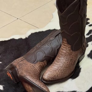 Op de afbeelding: Een paar bruine cowboylaarzen. Het onderste deel van de laarzen heeft een slangenpatroon, terwijl het bovenste deel van donkerder bruin leer is. De laarzen staan op een zwart-wit koeienvachtkleed.
