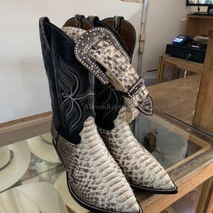 May include: A pair of black and beige snakeskin-patterned cowboy boots with pointed toes and intricate stitching on the shaft. A matching snakeskin-patterned belt with a large, ornate, crystal-encrusted silver buckle is draped over the boots.