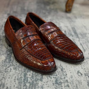 May include: A pair of brown leather loafers with a crocodile skin texture. The shoes have a classic design with a rounded toe and a low heel. The image is taken from a slightly elevated angle, showcasing the details of the leather.
