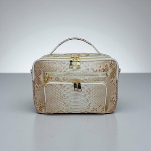 May include: A beige and white snakeskin-patterned handbag with gold-colored zippers and hardware. The bag has a top handle, a front pocket, and a shoulder strap. The bag is rectangular and appears to be made of leather.