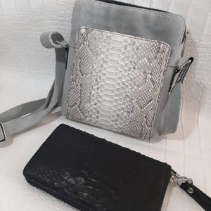 May include: A gray suede crossbody bag with a white and gray snakeskin front pocket. A black snakeskin wallet with a zipper closure is in the foreground. The bag has a shoulder strap.