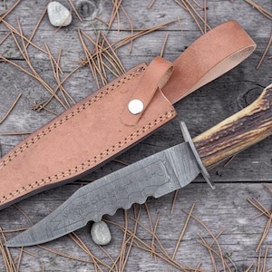 May include: A hunting knife with a patterned blade and a handle made of antler. The knife is accompanied by a brown leather sheath with a strap and a button closure. The knife and sheath are on a wooden surface.