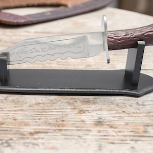 May include: A decorative knife with a serrated blade and a brown handle, displayed on a black stand. The blade has etched details. The knife is made of metal and has a silver finish.