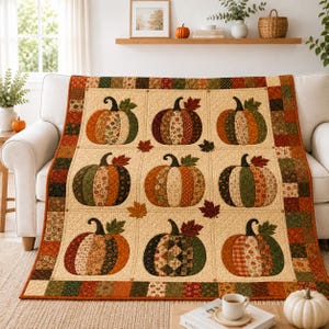Op de afbeelding: Een gewatteerde sprei met een raster van negen pompoenmotieven, elk gemaakt van verschillende stoffen met patronen in herfstkleuren. De quilt heeft een crèmekleurige achtergrond met kleine geborduurde bladaccenten en een patchwork-rand.