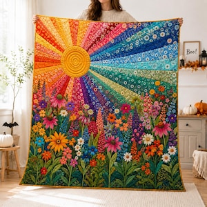 Op de afbeelding: Een rechthoekige quilt met een levendig zonnestraalpatroon in het bovenste gedeelte met stralende banen in geel, oranje, rood, blauw en groen. Het onderste gedeelte toont een dichte tuin met kleurrijke wilde bloemen, waaronder madeliefjes.