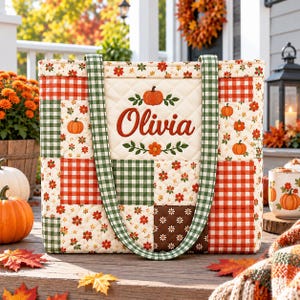 May include: A quilted tote bag featuring a patchwork design of red and green gingham, floral patterns, and brown squares. The center panel is cream-colored with a pumpkin graphic and the name Olivia printed in a dark orange script font.