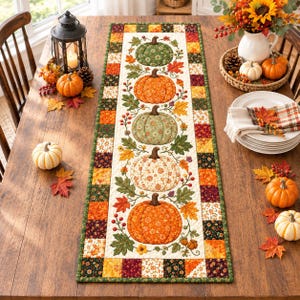 Puede incluir: Un camino de mesa rectangular de tela que presenta una fila vertical central de cinco calabazas estampadas en tonos verdes, naranjas y blancos, rodeadas de hojas de otoño y bayas, con un borde de pequeños cuadrados de patchwork multicolor.