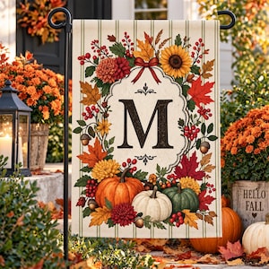 Könnte beinhalten: Eine rechteckige Gartenflagge mit cremefarbenem Hintergrund und vertikalen Streifen. In der Mitte befindet sich ein großer schwarzer Buchstabe M in einem dekorativen Rahmen, umgeben von Herbstlaub, Sonnenblumen, roten Beeren und Kürbissen.