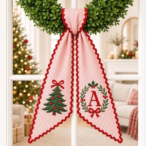 Può includere: Un decorativo appendiporta in tessuto con motivo a righe rosse e bianche e bordo smerlato rosso. Il lato sinistro mostra un albero di Natale verde ricamato con decorazioni rosse, mentre il lato destro presenta una lettera A rossa in una ghirlanda.