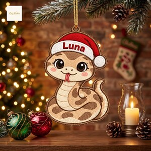 May include: A flat, embroidered Christmas ornament shaped like a cartoon snake with brown spots, a light tan belly, and a red Santa hat featuring the name Luna in white text. The ornament hangs from a gold string against a festive holiday backdrop.