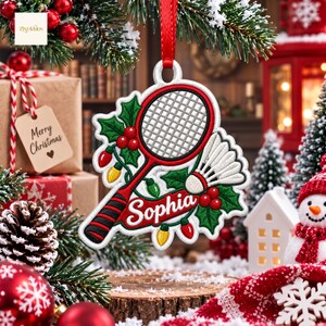 May include: A white embroidered Christmas ornament featuring a badminton racket and shuttlecock, decorated with green holly leaves, red berries, and colorful Christmas lights. A red banner across the center displays the name Sophia in white script.