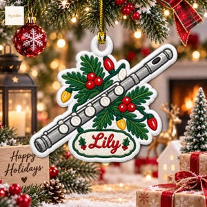 Peut inclure: Une décoration de Noël brodée blanche représentant une flûte argentée, des branches de pin vertes, des baies rouges et des lumières de fête colorées. Une étiquette blanche affiche le prénom Lily en rouge, orné de deux feuilles de houx.