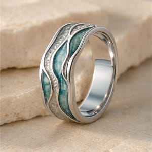 May include: A silver-toned metal ring featuring a wide band with a wavy, recessed design. The surface is inlaid with alternating sections of textured, shimmering silver-colored material and smooth, marbled teal-blue enamel in a flowing pattern.