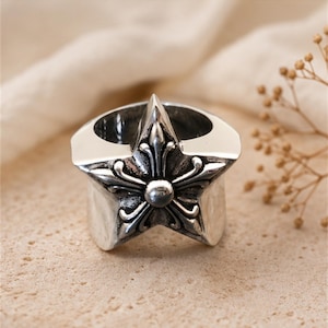 May include: A silver-colored ring with a star-shaped design. The star features a central floral element and ornate detailing. The ring has a wide band and a polished finish, suitable for everyday wear.