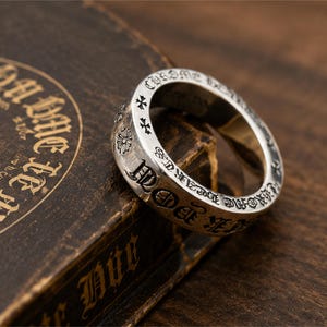 Peut inclure: Une bague en argent avec des inscriptions gravées et des motifs de croix et de fleurs. La bague est posée sur une surface sombre et texturée. L'inscription de la bague comprend les mots "CHROME HEARTS".