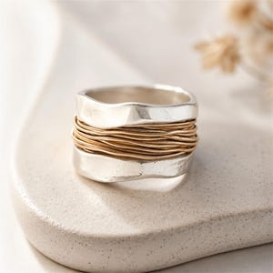 May include: A wide band ring featuring two thick, wavy-edged silver bands at the top and bottom, with a center section composed of multiple thin, coiled gold-toned wires wrapped horizontally around the circumference of the ring.