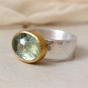 May include: A ring featuring a large, oval-shaped, faceted pale green gemstone set in a textured gold-toned bezel. The band is made of wide, hammered silver-toned metal with a reflective, uneven surface that catches the light across its width.