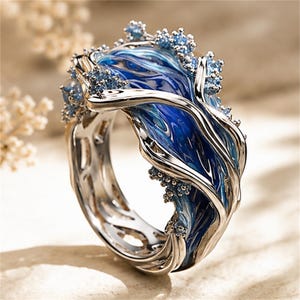 May include: A silver-toned ring featuring a swirling, deep blue enamel inlay that mimics the appearance of ocean waves. The band is accented with small, light blue crystalline clusters along the edges, creating a textured, organic design.
