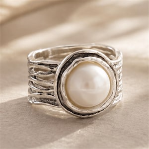 May include: A silver-toned ring featuring a large, round, white pearl set in a textured, organic-looking circular frame. The band of the ring is wide and composed of multiple layered, irregular, horizontal silver strands with a dark, oxidized finish.