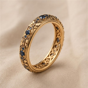 Può includere: Un anello a fascia in oro con intricati intagli floreali e piccole pietre preziose rotonde blu scuro incastonate sulla superficie esterna. La lavorazione del metallo presenta un motivo dettagliato di foglie e viticci con un design traforato.