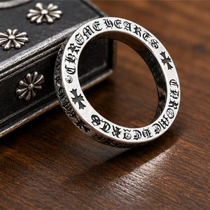 May include: A silver ring with the words "CHROME HEARTS" engraved around the outer edge. The ring also features cross designs. The ring is displayed on a wooden surface.