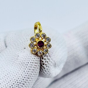 Puede incluir: Un anillo de nariz dorado con un diseño floral. El centro presenta una piedra preciosa redonda, de color rojo oscuro, rodeada por un anillo de piedras claras y brillantes. El anillo se sostiene sobre un fondo blanco texturizado.