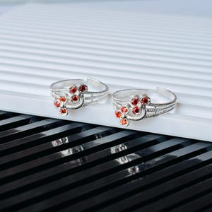 May include: A pair of silver-toned open-ended toe rings featuring a decorative floral motif. Each ring is adorned with three small, round, vibrant red stones arranged in a cluster, set against a textured, grooved band with an open back design.