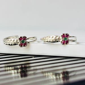 Op de afbeelding: Twee zilverkleurige ringen met een bloemachtig ontwerp. Elke ring heeft een centrale groep van vier kleine rode steentjes rondom één groen steentje. De band van elke ring is voorzien van een gestructureerd, bladachtig patroon aan de zijkanten.
