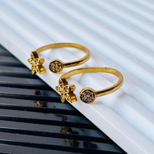 May include: Two gold-toned open rings featuring a five-petaled flower motif on one end and a small circular disc on the other. Both the flower petals and the circular discs are encrusted with small, clear, sparkling stones throughout the surface.
